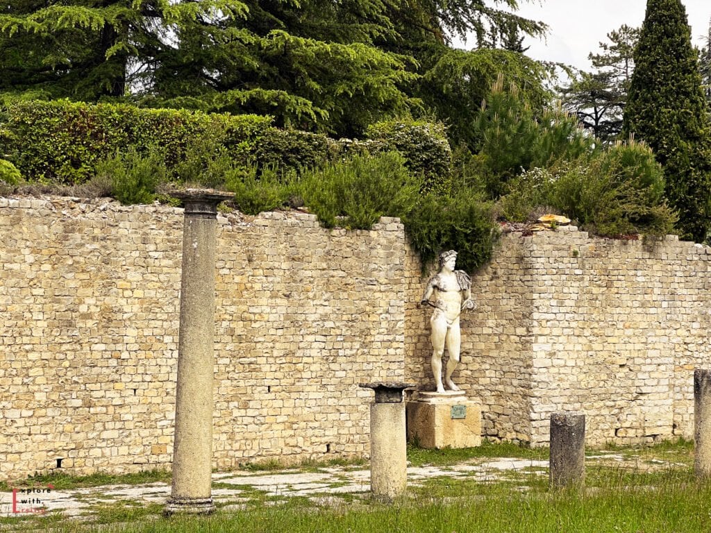A white marble statue of a muscular male figure, possibly Hercules, stands against a stone wall in the ancient ruins of Vaison-la-Romaine, France. Surrounding the statue are remnants of Roman columns and lush greenery atop the wall, with tall evergreens in the background.